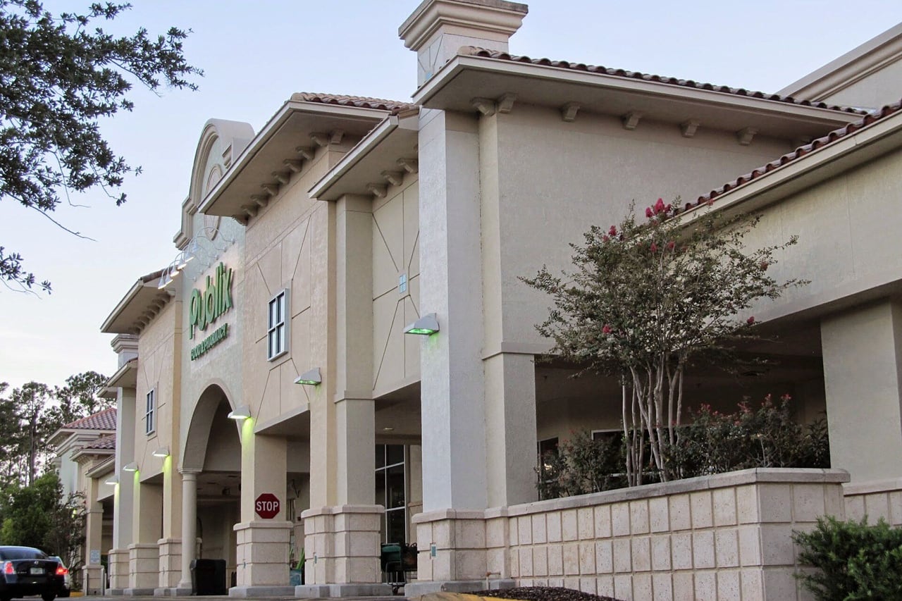 Fachada creme do supermercado Publix com arcos, telhado de telha e árvore florida na frente.